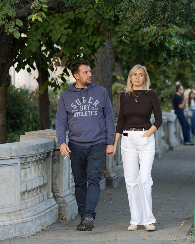 A man wearing a Superdry blue hoodie and dark trousers holding hands with a woman in a dark long-sleeve top and white wide-leg trousers while walking along a sidewalk
